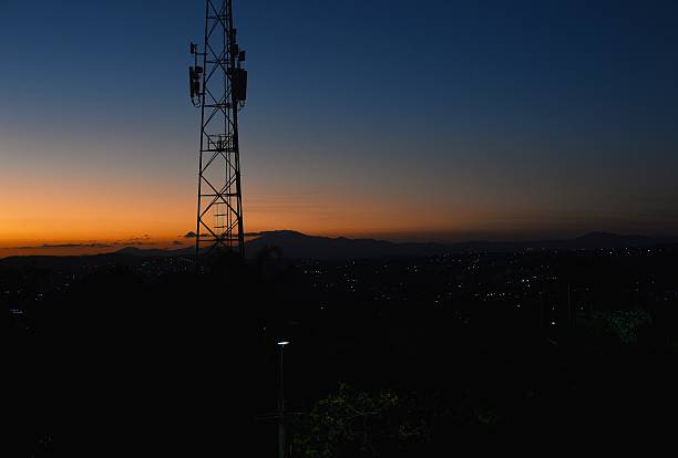 A telecommunications tower stands in silhouette against a vibrant sunset over a Brazilian hillside town. The warm orange tones fade into deep blue as twilight sets in, revealing a scattering of city lights below. The image captures the contrast between nature and technology in a tranquil evening landscape, with soft contours of the mountains in the background.