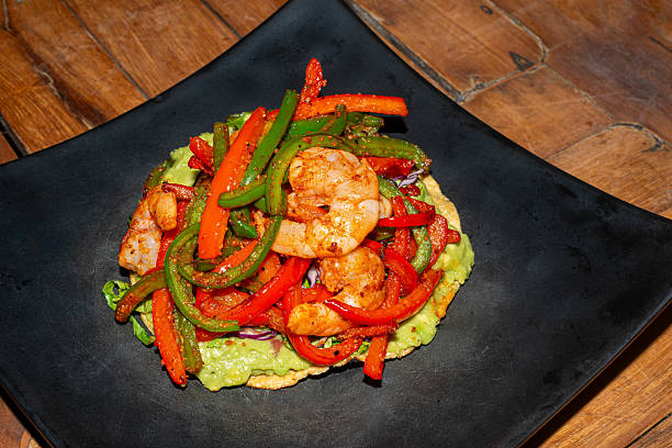 Close-up of a traditional Mexican shrimp tostada served with guacamole and peppers on a black plate over a rustic wooden table. A colorful and delicious seafood dish that represents authentic Mexican cuisine.