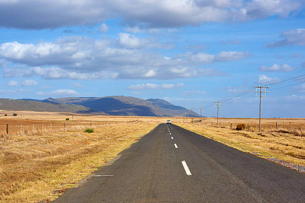 An expansive view of a road in Western Cape, South Africa, bordered by serene farmland and hills in the background, under a vivid blue sky adorned with scattered clouds on a sunny day.