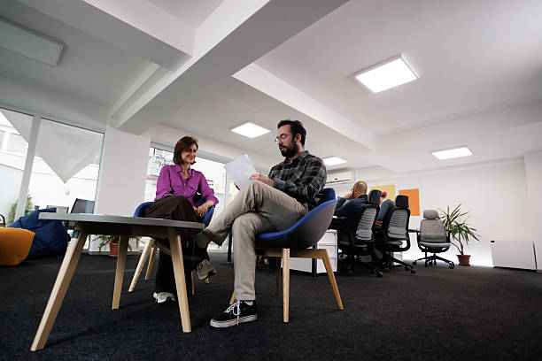 Two businesspeople engage in a discussion while seated in an open office area filled with plants and informal seating. Colleagues are seen working in the background.