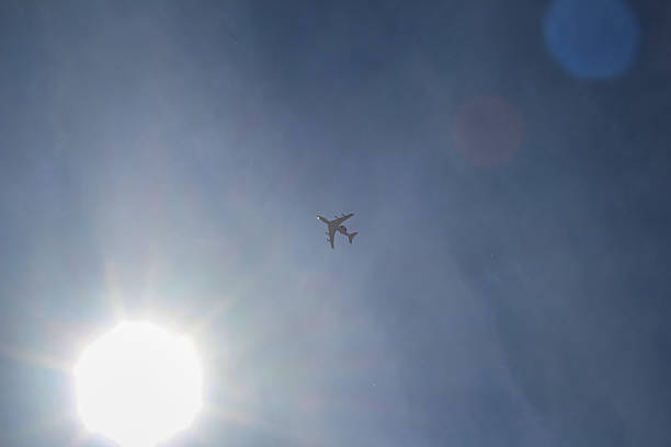 The silhouette of a passenger airplane is clearly visible in the blue sky, flying high near the bright sun. Light flares and the sun's halo add drama to the image