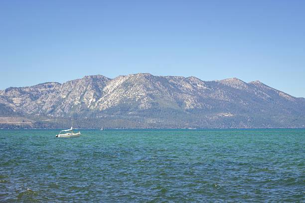 This image captures a calming scene of a small sailboat floating against the majestic mountains of Lake Tahoe. The vivid shades of blue water and green mountains create a perfect blend for relaxation and enjoyment of nature. This location is popular among tourists seeking solitude and the beauty of untouched nature