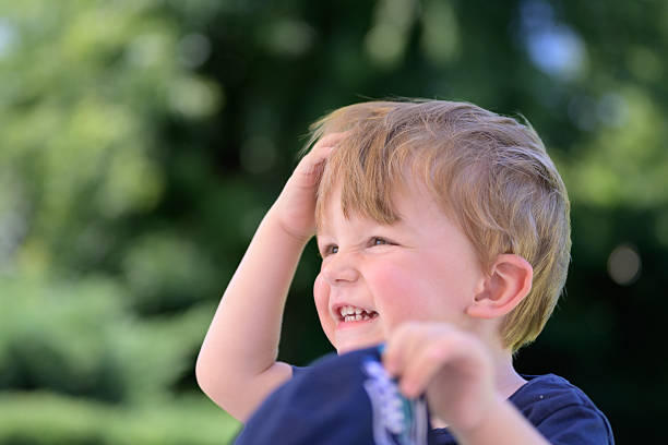 A cheerful young boy laughs joyfully while touching his hair outdoors on a sunny day.