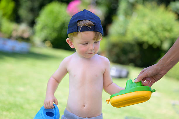 A toddler in a cap holds a colorful toy boat handed by an adult while carrying a blue watering can.