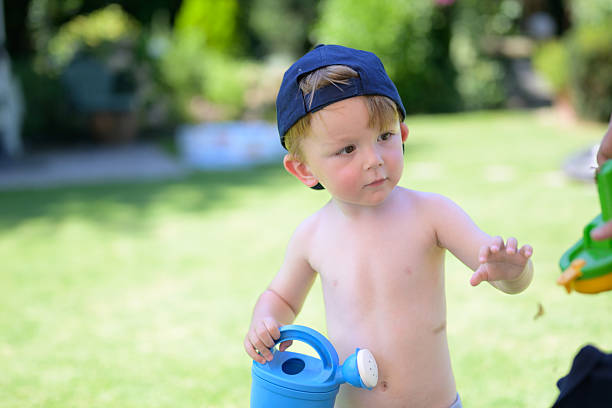 A toddler with a cap holds a blue watering can and reaches out towards a colorful toy outdoors.