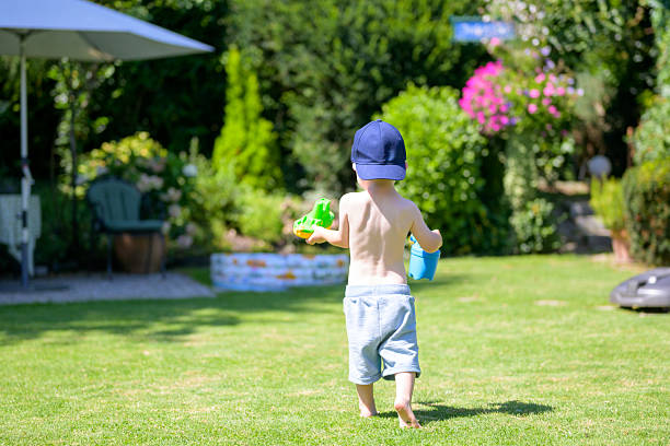 A young child in a cap plays barefoot on the grass carrying toys in a sunny garden.