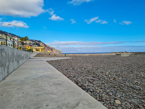 Wide-angle view of the seaside promenade and pebble beach in Funchal, Madeira, Portugal, likely Praia Formosa. The foreground features a wide concrete walkway that borders a vast stretch of dark, volcanic pebbles and stones.