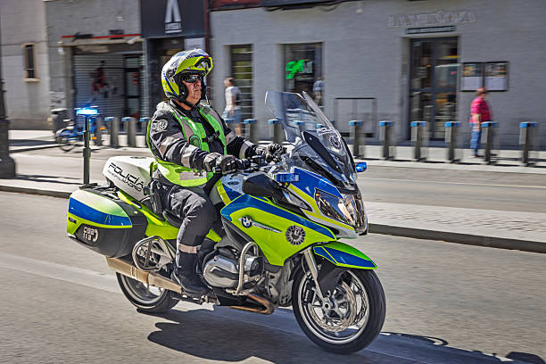 Madrid, Spain, Europe, September 12, 2025: Dynamic urban scene in central Madrid featuring a Spanish police officer riding a high-visibility motorcycle while escorting a vehicle through city traffic. The officer wears a helmet and reflective gear, navigating a tree-lined street bordered by modern buildings and a glass bus shelter. Pedestrians and parked cars frame the background, adding depth to the metropolitan setting. The vibrant colors of the police motorcycle stand out against the subdued tones of the urban environment.