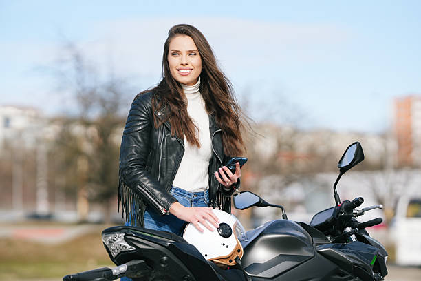 A young woman flashing a bright smile while seated on a motorcycle, showcasing her trendy leather jacket and interacting with her mobile phone. This image captures modern lifestyle habits and symbolizes freedom, empowerment, and the joy of being on the road, infused with a contemporary vibe.