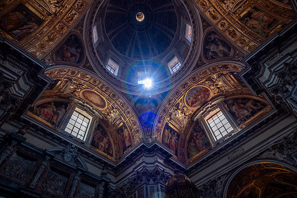 Light beams shine through a grand cathedral dome, revealing intricate Baroque frescoes, windows, and sacred religious architectural details.