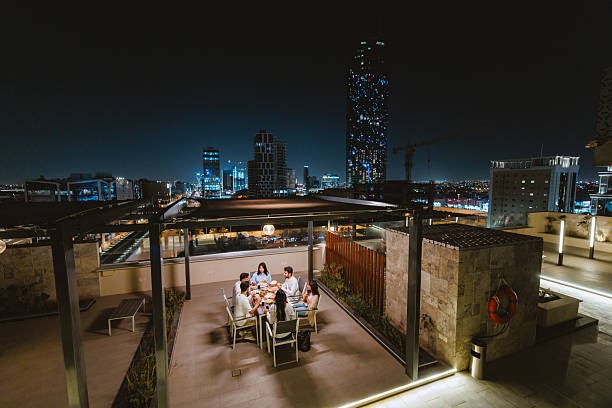 Young Arabic friends dining on a rooftop terrace in Riyadh featuring a stunning night cityscape. The group appears cheerful and relaxed, enjoying a meal together under ambient lighting with a view of the skyline.