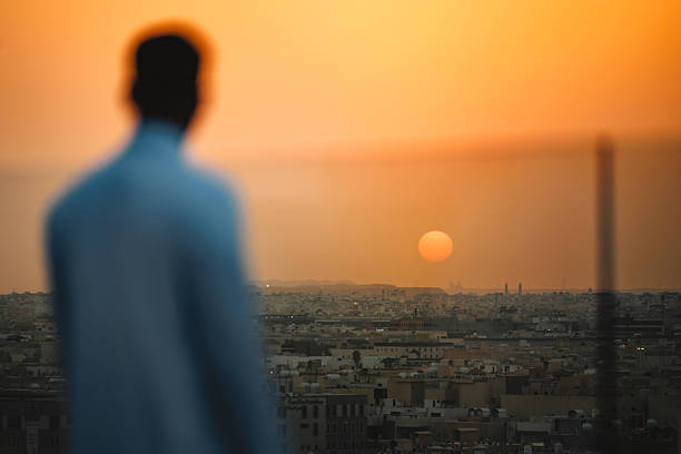 Portrait of a young Arabic male enjoying a serene sunset view over Riyadh from a rooftop terrace featuring the city's expansive landscape and vibrant orange sky.