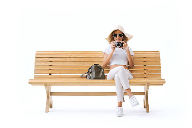 Happy female tourist sitting on a wooden bench and checking her camera, isolated on white background