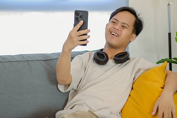 istock Young Man with Down Syndrome Holding Mobile Phone Doing Video Call on a Couch 2238714054