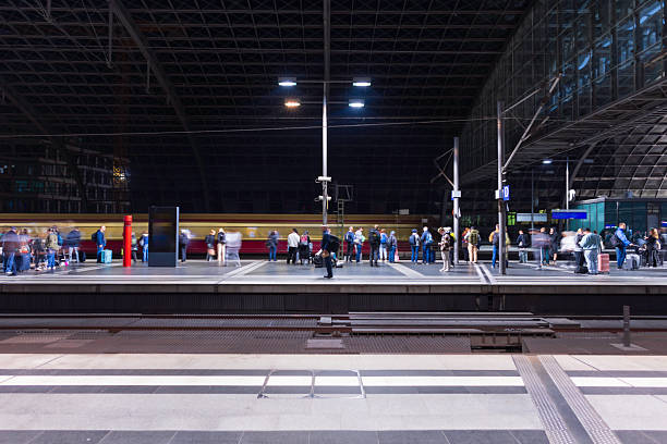 A crowded train platform at night with a red train passing by in the background. Modern glass and steel architecture overhead, with travelers waiting, moving, and luggage in motion blur.
