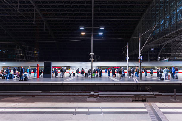 A busy, modern train platform at night. Passengers stand in line with luggage as a fast train passes by, illuminated by bright lights, capturing urban travel and transit life.