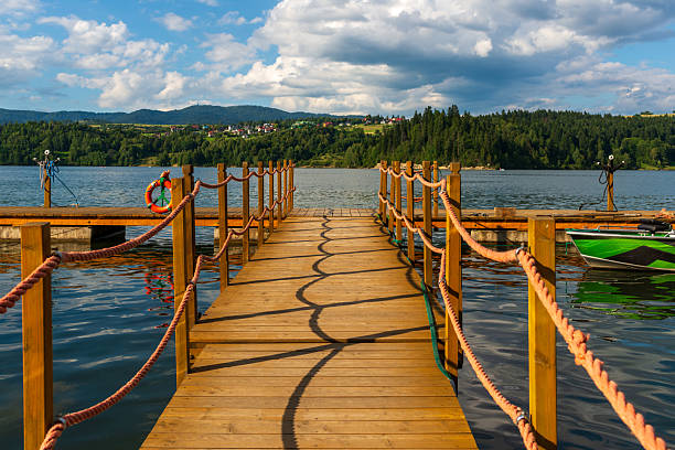 A wooden pier with orange rope railings casts rhythmic curved shadows on the water surface. Clear sky with fluffy clouds and forested hills create a calm, inviting summer lakeside scene.