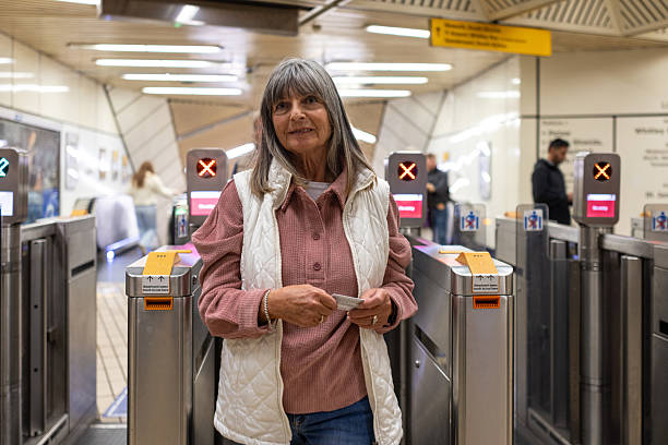 Medium shot of a senior adult female walking through ticket barriers at a metro station in North East England. She is holding a ticket in both hands, wearing casual clothing.\n\nVideos similar to this scenario available.