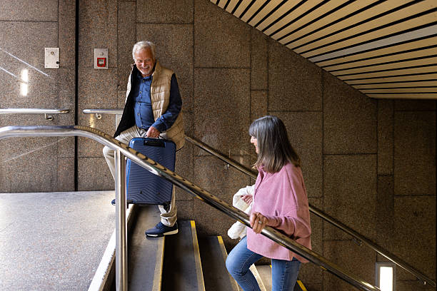 Medium side-view shot of a senior couple walking up the staircase of a metro station in North East England. They are both wearing casual clothing, carrying suitcases. The male is walking slightly ahead, turning to smile over his shoulder at his partner.Videos similar to this scenario available.