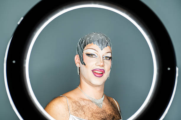 A latin drag queen poses confidently in front of a ring light, showcasing vibrant makeup and stylish jewelry. The background is softly lit, enhancing the striking features and creative look.