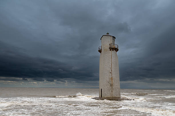 A lighthouse sits on a small rocky island on the horizon as it shines its light during inclement weather.