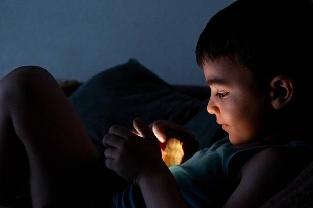 A boy is sitting on a bed and looking at a phone. The room is dark and the boy is the only person in the image