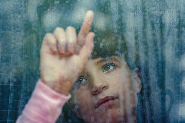 Child looking out rainy window with finger on glass stock photo