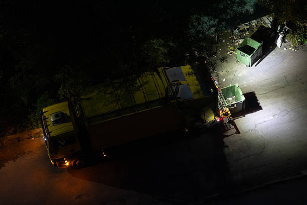 The garbage truck stands near trash containers on the side of a city street at night. The truck's hazard lights and taillights are on. Two plastic trash containers are parked next to the truck. View from above.