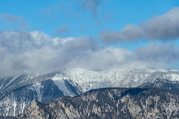 Majestic alpine mountains covered in snow with rugged rocky ridges and forested slopes. The dramatic clouds and blue sky create a scenic winter landscape, perfect for themes of travel, adventure, and natural beauty in the Alps.