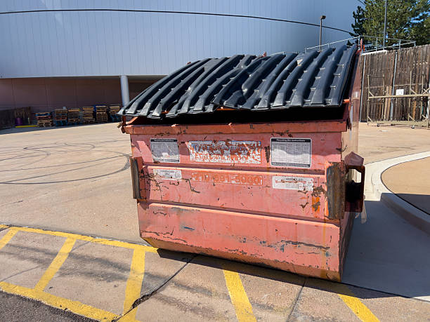 Parker, Colorado, USA-September 18, 2025 - A large rusty metal dumpster is positioned on a concrete surface in an outdoor parking area. The container is weathered with peeling paint and sits in front of a tall wooden fence under clear daylight. The surroundings include marked asphalt and part of an industrial building structure.