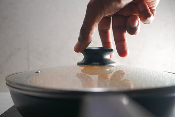 A hand is placing a lid on a pan atop a stovetop for cooking.