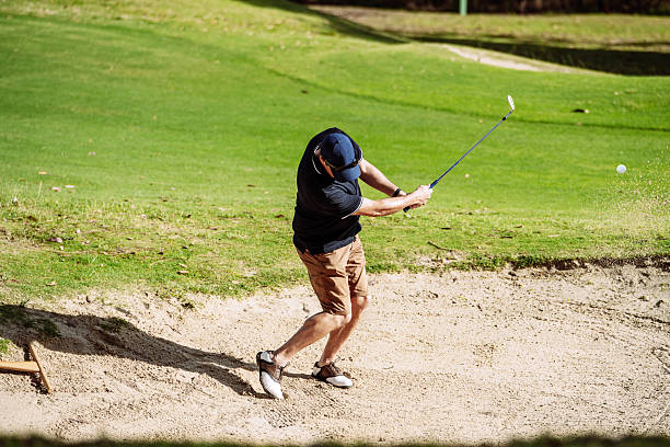 Male golfer performing a sand shot with follow through on a sunny course, illustrating skill and precision in a competitive match.