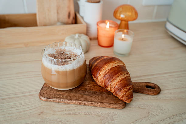 Cup of hot latte with foam, candle, croissant on the table. Autumn drink.