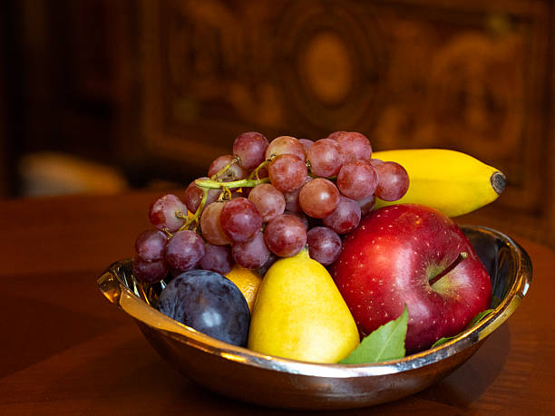 As seen in natural light, a red apple, yellow pear, purple plum, purple grapes, yellow banana and a hidden orange are served in a silver fruit bowl placed on a wooden table in a hotel room.  The ingredients offer people healthy eating food for a healthy lifestyle.