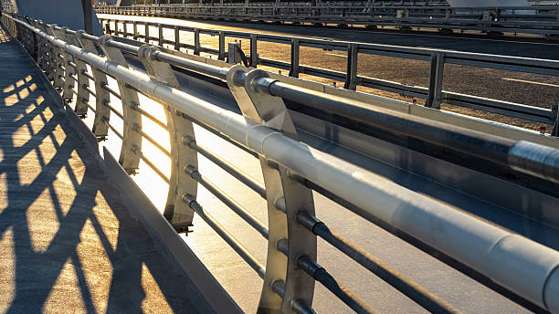 Bright sunlight casts long shadows on sleek metallic railings of a pedestrian bridge in the late afternoon.