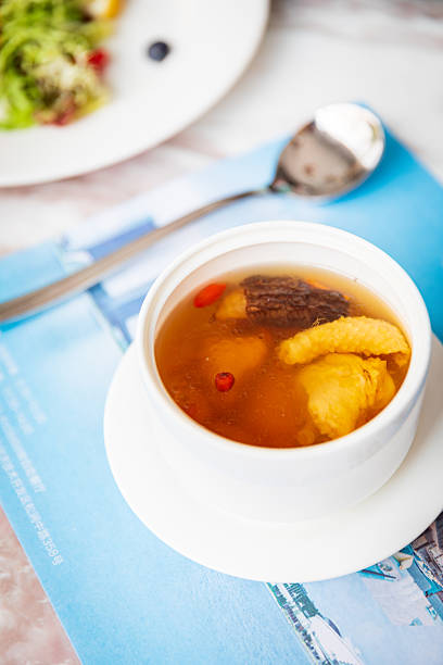 A bowl of exquisite soup, its clear broth dotted with tender, fragrant mushrooms and slow-cooked chicken. Blue-and-white porcelain pieces are arranged neatly on the pristine white tabletop, adding an authentic touch to the dining experience while accentuating the warmth and comfort this dish brings