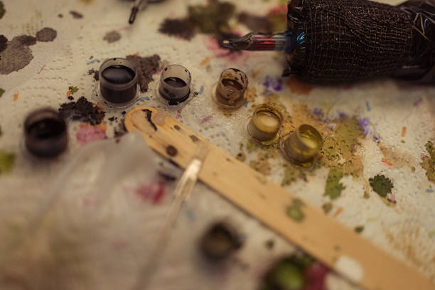 Close-up view of various small colorful tattoo ink cups and tattoo tools on a textured surface covered with ink stains and residues, highlighting the detailed preparation for tattooing in a professional setting.