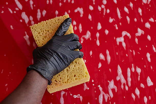 Black adult hand wearing black glove cleaning red surface with yellow sponge, creating soap suds and bubbles, close up showing detailed texture of sponge and skin