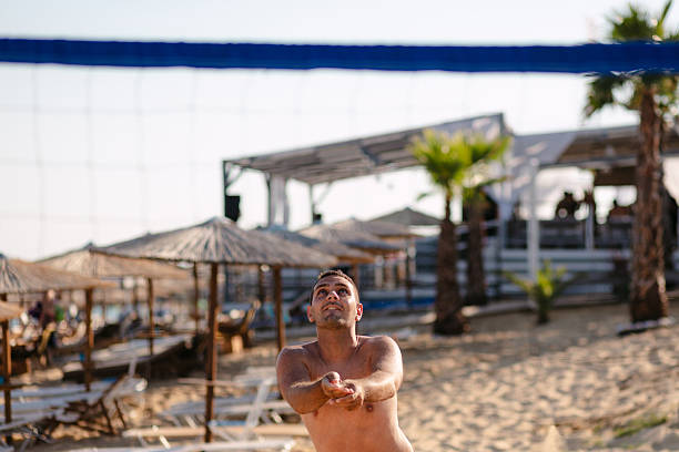 A shirtless male volleyball player in colorful shorts prepares to spike a ball on a sunny beach. Palm trees, cabanas, and a volleyball net create a lively, resort-style scene