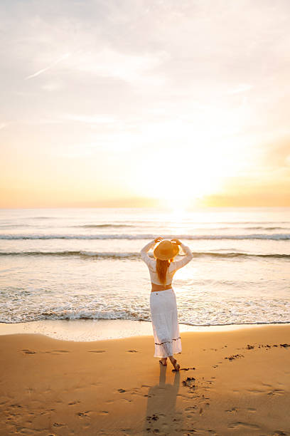 A young woman strolls along a sandy beach at sunrise. A beautiful woman in a hat enjoys the seascape, feeling free. Concept: vacation, weekend, and enjoyment.