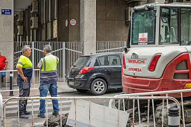 Madrid, Spain, Europe, September 10, 2025: Two male construction workers in high-visibility gear stand near a fenced-off urban worksite in Madrid, Spain, engaged in conversation about the ongoing roadwork. A red and white Takeuchi excavator labeled JOFEMESA is parked nearby, surrounded by metal barriers and a partially dug-up street. The workers’ clothing and body language suggest active coordination and planning. The backdrop includes a gray building facade with air conditioning units and a visible street sign, adding geographic and editorial context to the scene.