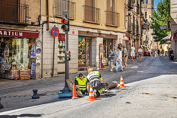 Toledo, Spain, Europe, September 16, 2025: Two male utility workers in high-visibility uniforms kneel beside an open utility hole on a cobblestone street in Toledo, Spain, handling tools and technical components. Surrounded by traffic cones and scattered equipment, the scene captures a moment of hands-on infrastructure maintenance in a busy urban environment.