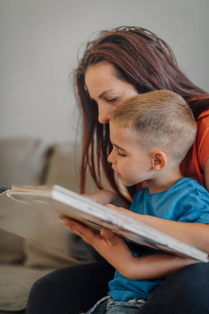 Young boy reading a book while lying on a sofa next to his smiling mother, enjoying a quiet moment of learning and bonding