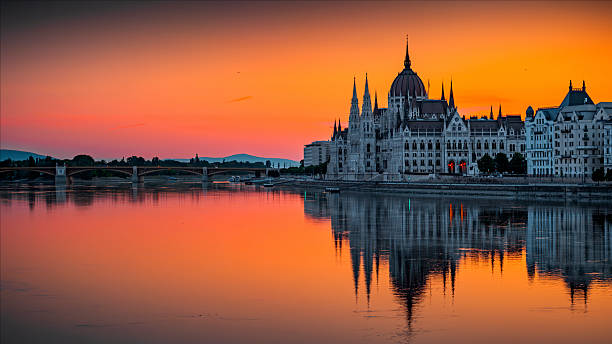Hungarian Parliament Building in Budapest, Hungary, during a vivid sunset. The Gothic Revival architecture is mirrored in the calm surface of the Danube River, with glowing orange and pink skies in the background. Budapest in Hungary - Eastern Europe