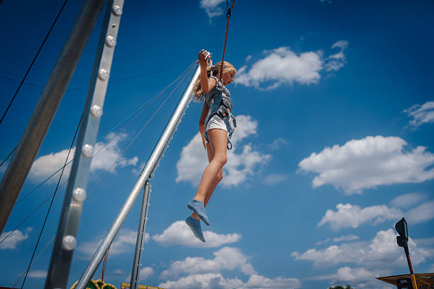 Girl wearing safety harness enjoying bungee jumping trampoline against blue sky with white clouds in amusement park during summer day