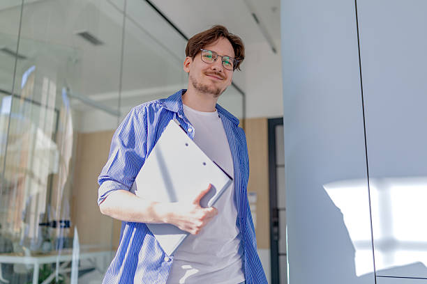 istock Young man smiles confidently while working in a modern office setting, holding a clipboard and engaging with his tasks 2238920923