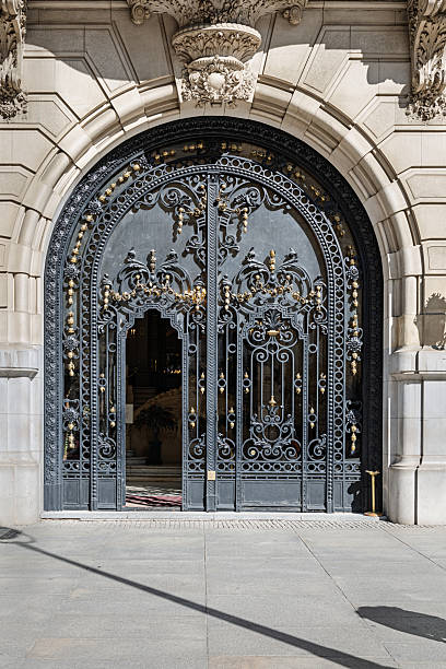An elaborately designed wrought iron gate stands beneath a carved stone archway in central Madrid, Spain. The gate features intricate scrollwork and floral motifs in black and gold, framed by classical stone detailing including a central crest and symmetrical embellishments. Partially open, the gate reveals a glimpse of the interior with patterned flooring and warm lighting. The image highlights the craftsmanship and grandeur of historic European architecture, blending decorative ironwork with monumental stone design in an urban setting.