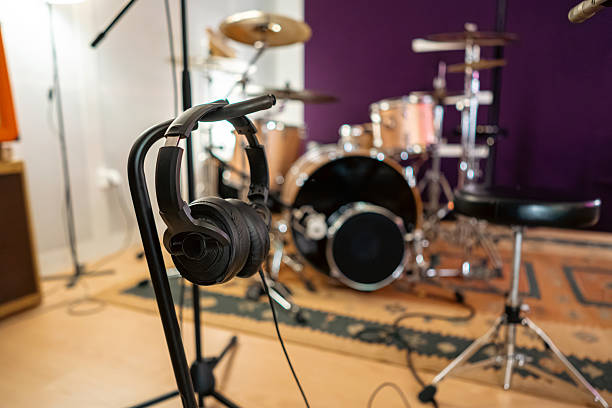 A pair of professional studio headphones placed in a recording studio environment during a soundcheck session. Surrounded by musical instruments like drums and acoustic guitars, and audio equipment including a microphone with a pop filter