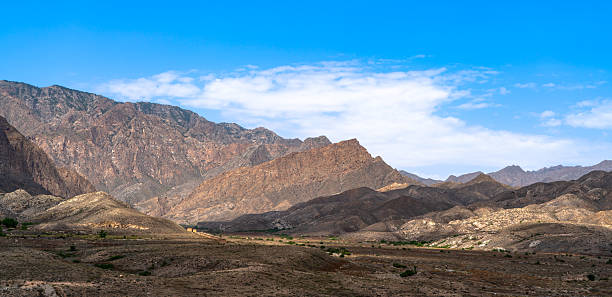 This dramatic landscape captures the imposing majesty of jagged, terraced red and brown rock formations rising in layered ridges against a brilliant azure sky scattered with soft white clouds, with a vast, sun-baked valley floor dotted by sparse green shrubs in the foreground. The warm earthy tones and expansive scale evoke raw wilderness adventure and timeless geological wonder, ideal for travel posters, outdoor exploration visuals, environmental documentaries, or inspirational nature themes in advertising, editorial, or lifestyle campaigns.