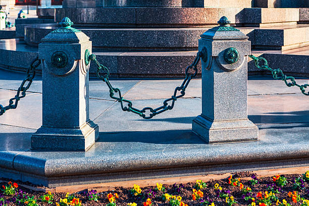 Granite bollards connected by a weathered, dark green chain barrier at the base of a monument in Vienna. Decorative stone bollards linked by chains stand before grand stone structure and flower bed